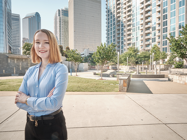 A student stands, arms crossed, in a light blue blouse and black slacks in a city park, surrounded by gleaming skyscrapers.